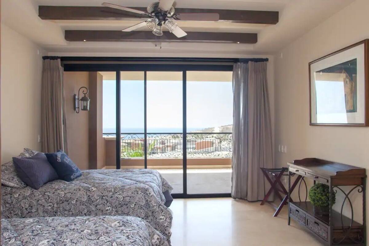A bedroom with a bed, a ceiling fan, a nightstand, a window with curtains, and a view of the outside.