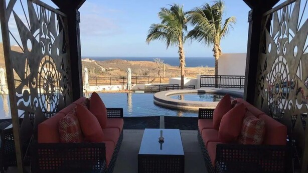 A terrace with two red sofas and a table, overlooking a pool and palm trees with a view of the coastline. - CONDO Q BY TRVL2HM (Cabo San Lucas)