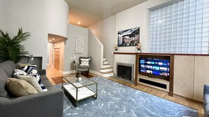 A modern living room with a grey sofa, a glass coffee table, a fireplace, and a large TV screen.