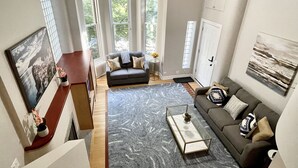 A spacious living room with a grey sofa, a coffee table, and a large rug. There are three windows providing natural light and a view of greenery outside.