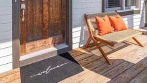 A house with a wooden front door, a welcome mat, a wicker chair with orange cushions, and a window with blinds. - Pet Friendly Large House | 10 Min to Broadway (Nashville)