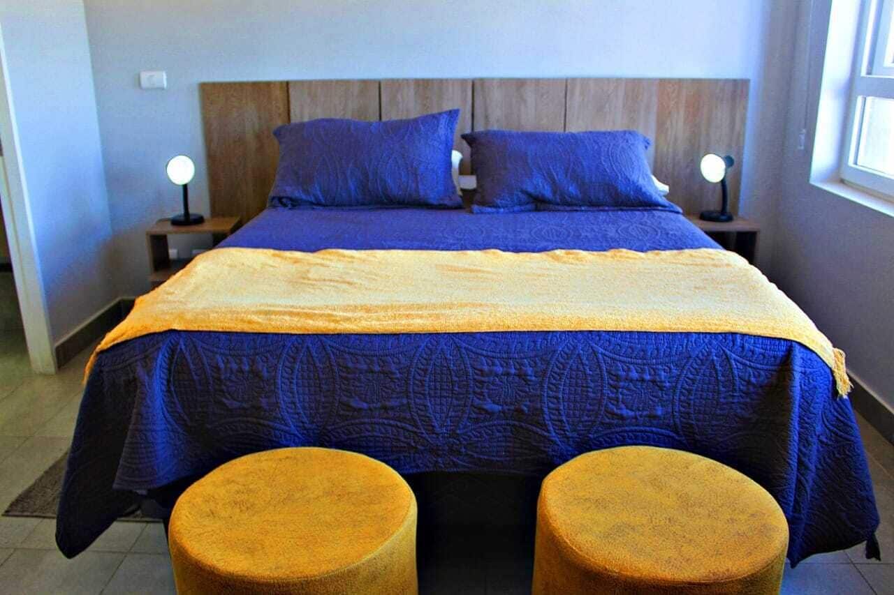 A bed with a blue and yellow bedspread, two pillows, two bedside lamps, and two round ottomans.