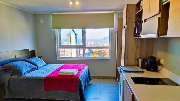 A compact room with a bed, a small kitchenette, and a window with a city view. - Apartamentos Bauerle (Temuco)