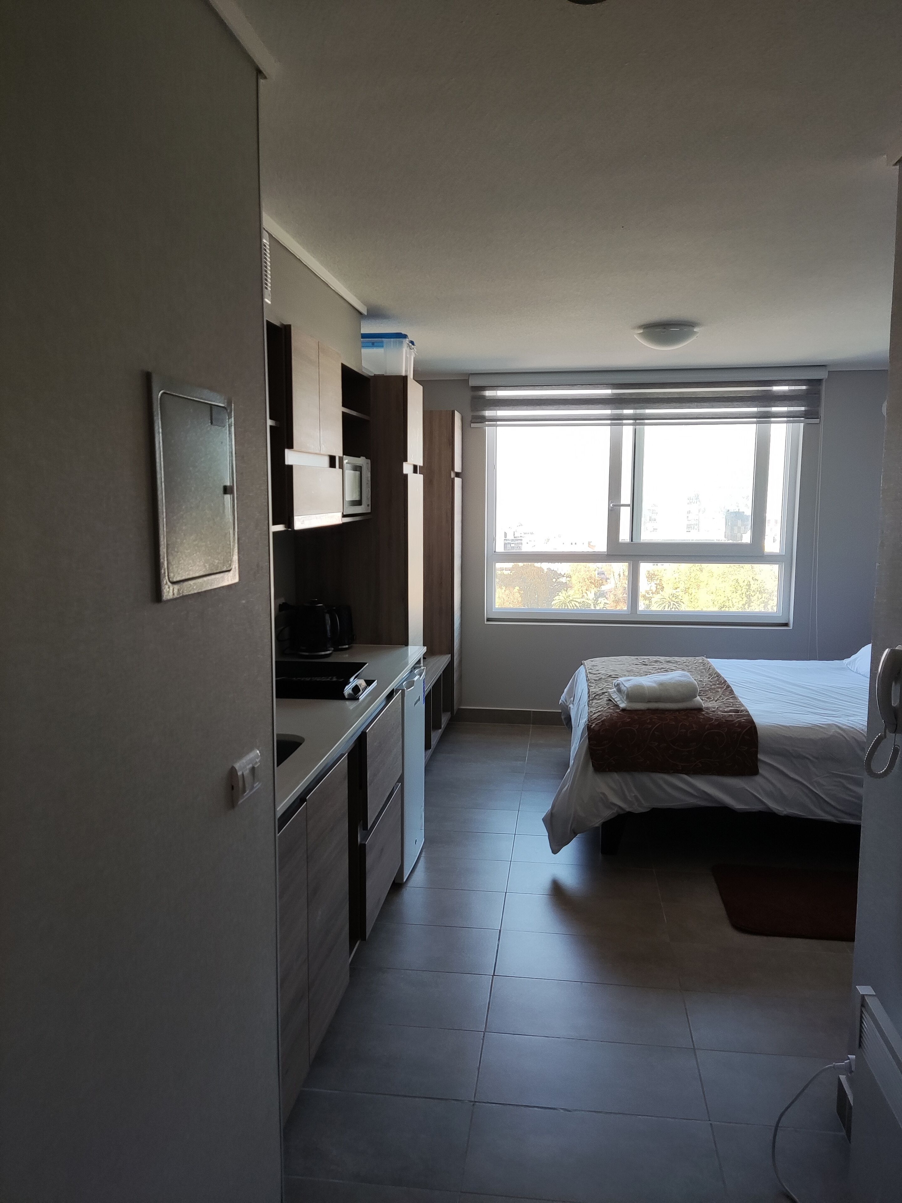 A compact hotel room with a kitchenette, a bed with a patterned bedspread, and a large window providing a view of the outside.