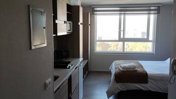 A compact hotel room with a kitchenette, a bed with a patterned bedspread, and a large window providing a view of the outside.