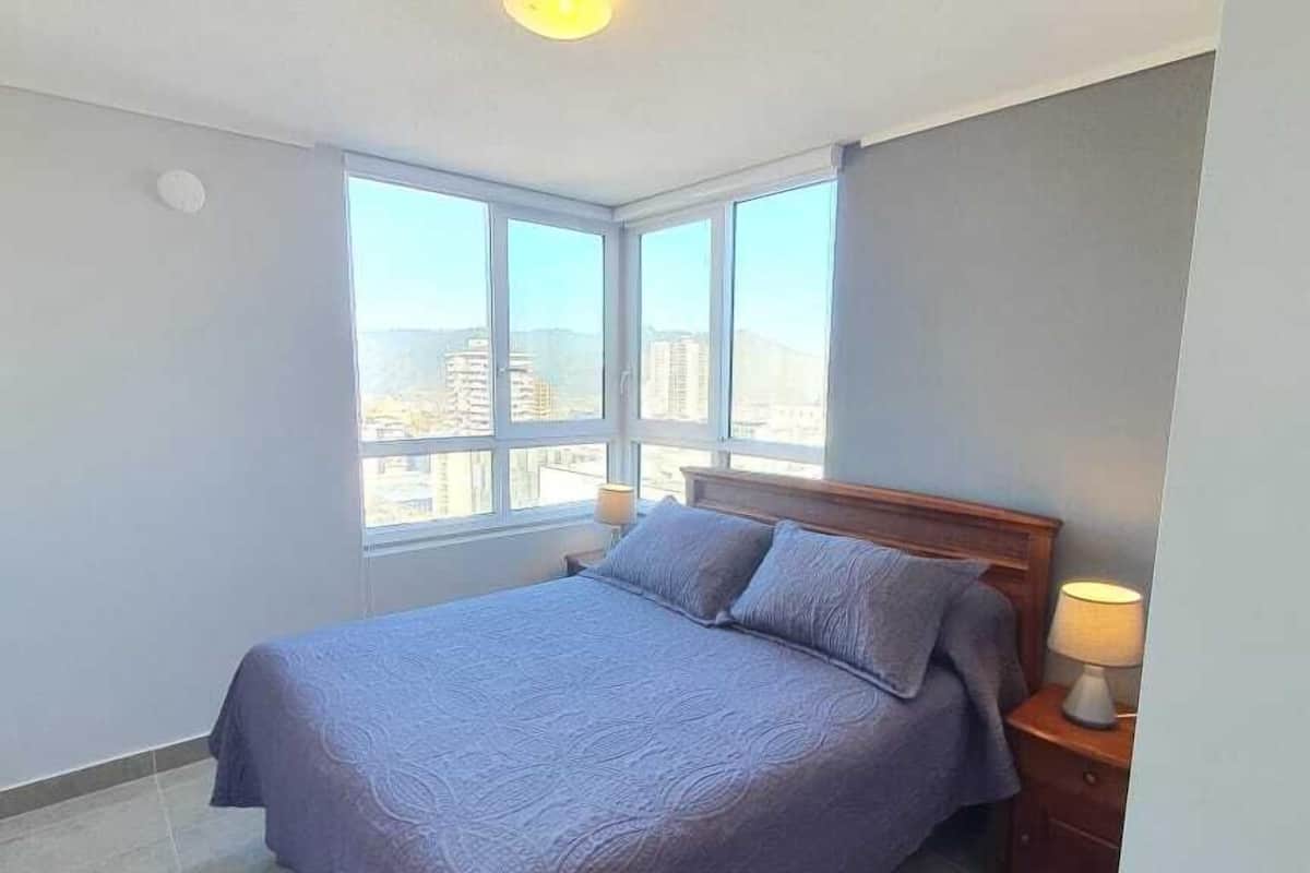 Un dormitorio con una cama grande, una mesita de noche, una lámpara y vistas amplias de la ciudad a través de grandes ventanales.