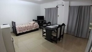 A small, well-lit room with a bed, a desk with a chair, a television, and a window with curtains.