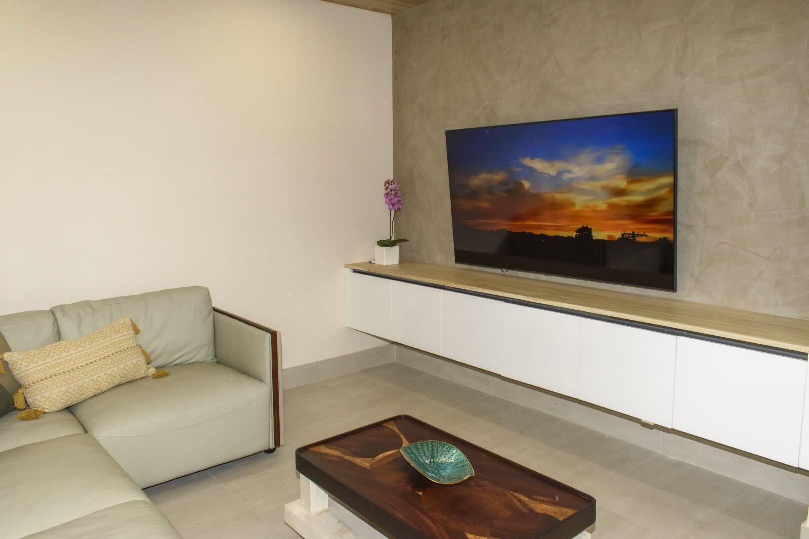 A modern living room with a light-colored sofa, a wooden coffee table, and a mounted TV displaying a sunset scene.