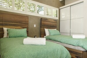 A hotel room with two beds, each with green bedding and white pillows. There is a wooden headboard, a wardrobe, and a window with blinds.
