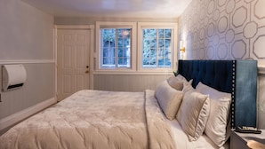 A bedroom with a large bed, a chandelier, a window with a view of trees, and a patterned wall. - Boho Manor Resort - Room B (Monte Rio)