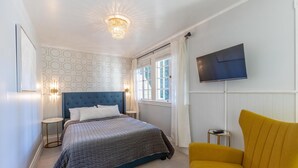 A bedroom with a bed, a television, a yellow armchair, and a window with curtains. - Boho Manor Resort - Room 28 (Monte Rio)