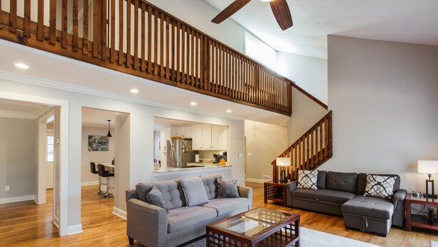 A spacious living room with hardwood floors, a gray sofa, a glass coffee table, and a staircase with wooden railings.