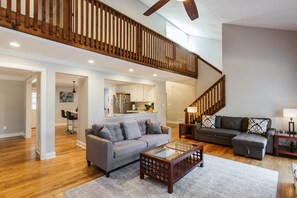 A spacious living room with hardwood floors, a gray sofa, a glass coffee table, and a staircase with wooden railings. - A Spacious Place for Everyone. Sanford Stadium, Downtown and Dining So Close! (Athens)