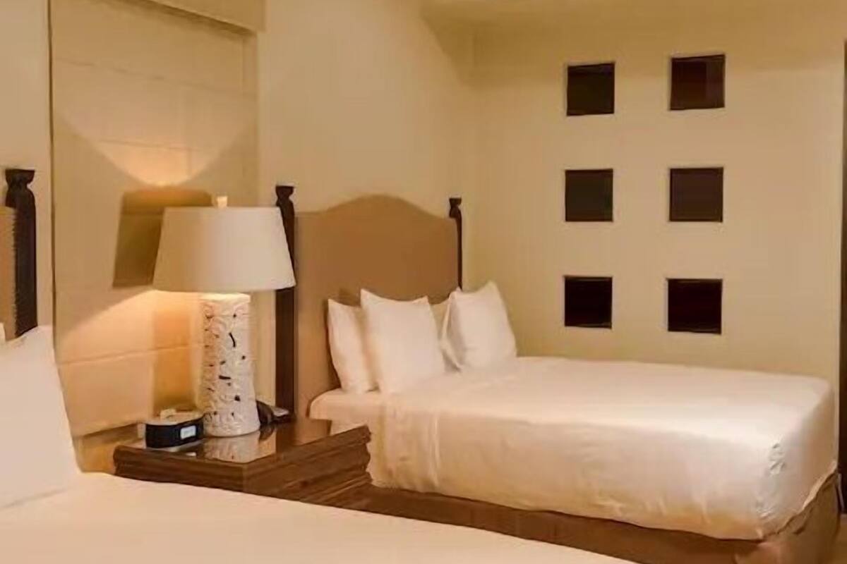 A hotel room with two beds, a ceiling fan, a nightstand with a lamp, and a wall with square cutouts.