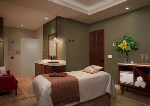 Une chambre moderne avec un lit, une coiffeuse avec un miroir, un fauteuil rouge, un meuble en bois contenant des serviettes et un vase avec des fleurs.