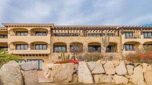A multi-story hotel with a stone facade, arched balconies, and a courtyard with cacti and rocks. - CONDO A BY TRVL2HM (Cabo San Lucas)
