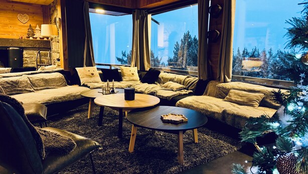 Ein gemütliches Wohnzimmer mit einem Ecksofa, einem Couchtisch und einem Kamin. Große Fenster bieten Blick auf Bäume und Gebäude.