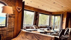 Ein gemütliches Blockhaus-Interieur mit Steinwand, Holzboden und großen Fenstern mit Blick ins Freie.
