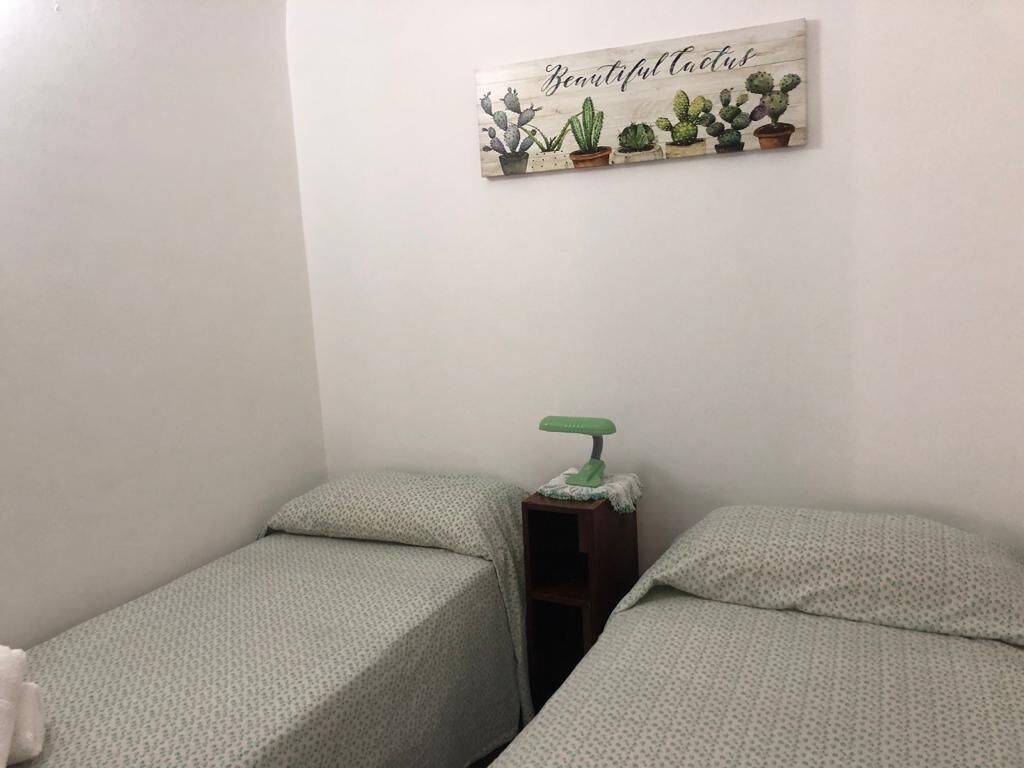 Quarto pequeno com duas camas, uma mesa de cabeceira com abajur e uma decoração de cacto na parede.
