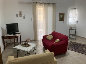 Een woonkamer met een rode bank, een beige bank, een glazen salontafel met een bloemen decoratie, een televisie op een houten tafel, en een glazen plank met decoratieve objecten.