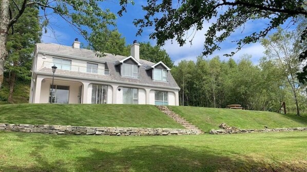 A two-story house with a stone retaining wall, a grassy lawn, and a wooded backdrop.