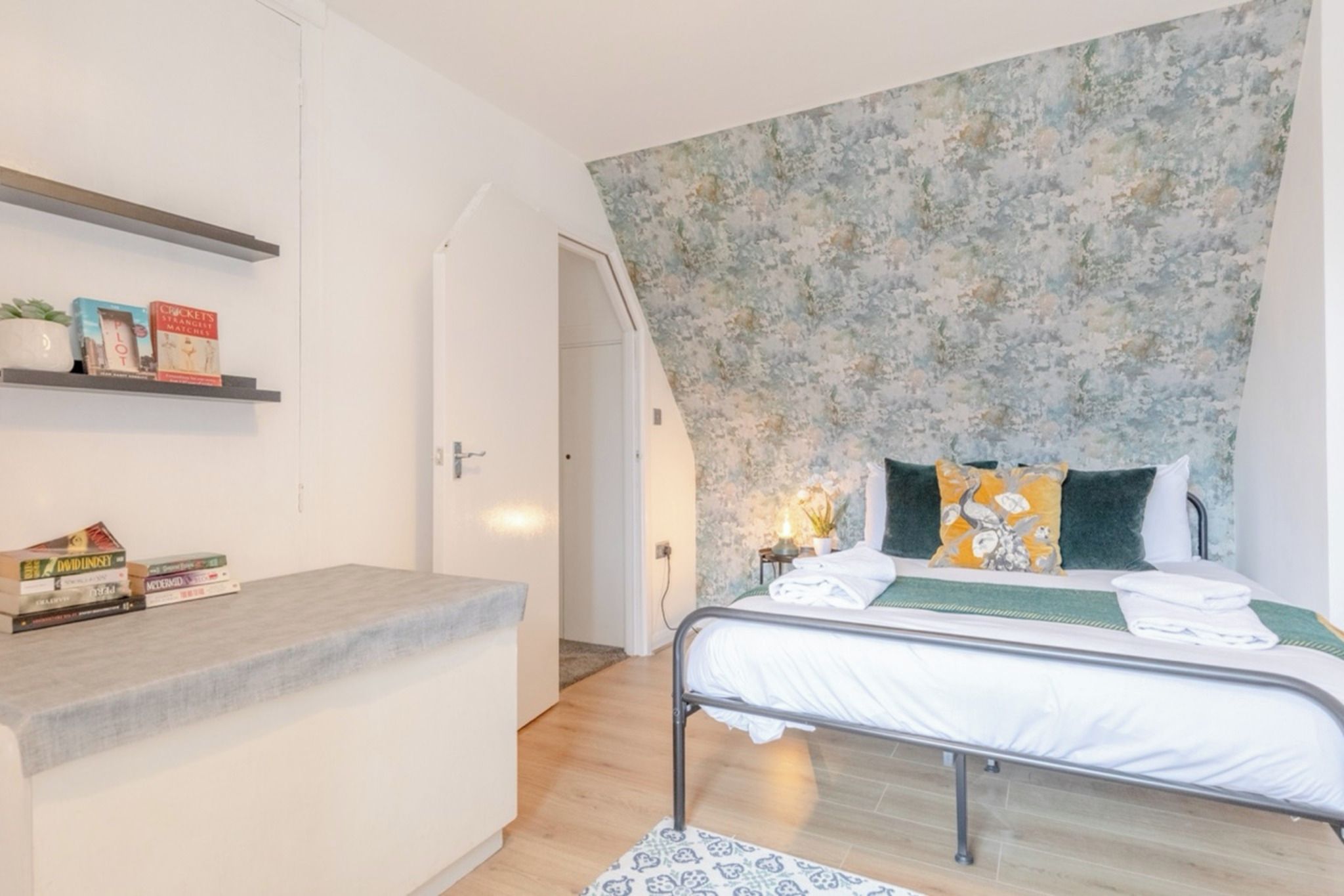 Een slaapkamer met een bed, nachtkastje en een plank met boeken.