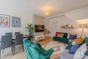 A modern living room with a dining area, a flat-screen TV, a sofa with colorful cushions, and a large plant. - Central 3BD House With Garden - 4min walk to Tube! (London)