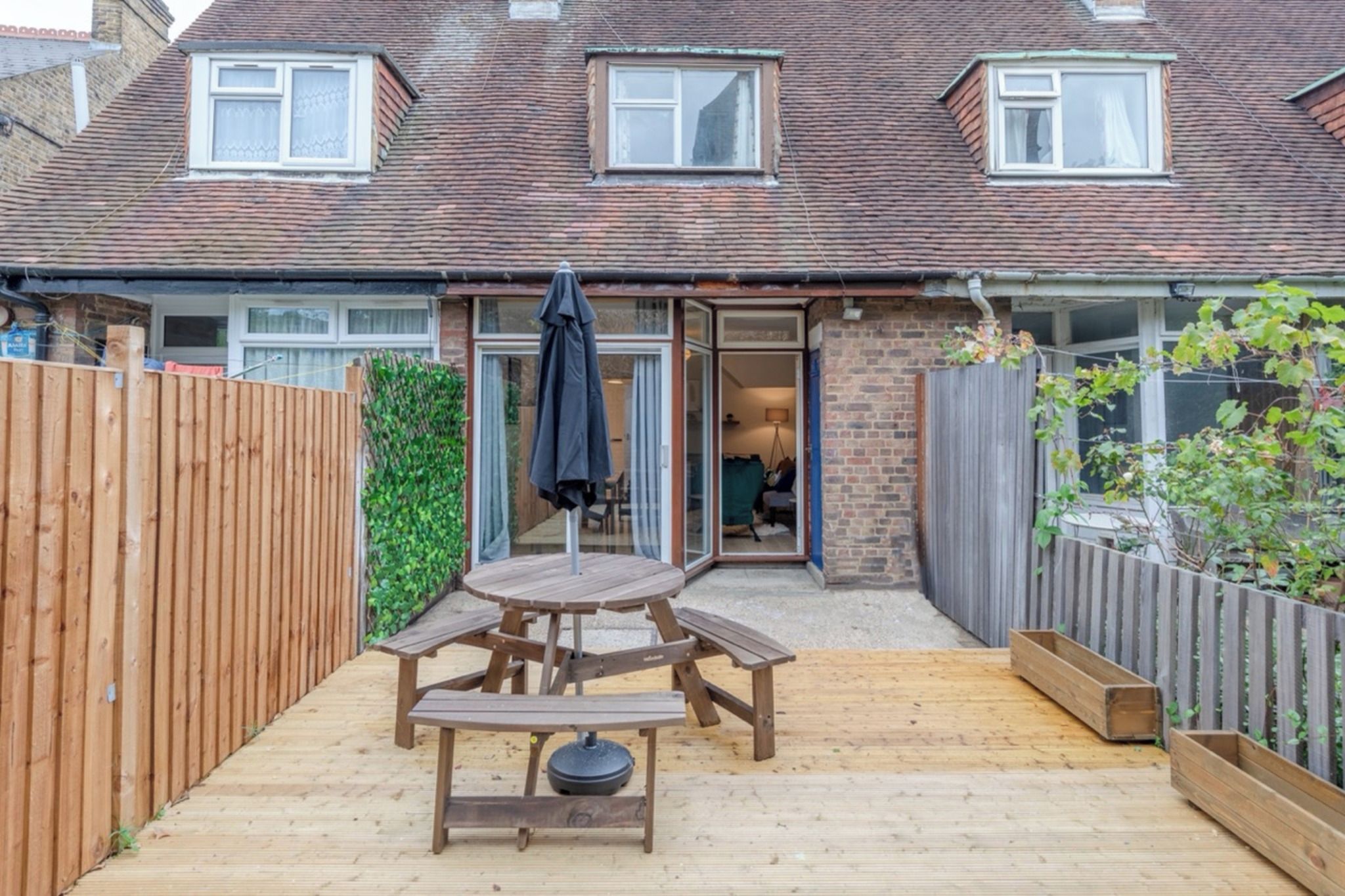 Une terrasse en bois avec une table de pique-nique et des bancs, entourée d’une clôture en bois et d’une maison en briques avec un toit en tuiles.