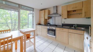 A modern kitchen with wooden cabinets, a gas stove, an oven, and a dishwasher. There is a dining table with chairs and a window with a view of trees. - Spacious 2BDR Flat in the Heart of West London (London)