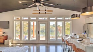 A modern kitchen with a large island, bar stools, and a view of the outdoors through large windows. - Modern Norfork House w/ Patio on White River! (Norfork)