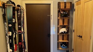 A hallway with a wooden door, skis and snowboards on the left, and a shelf with helmets and bags on the right. - Okemo Condo on Bus Route in Center of Town (Ludlow)