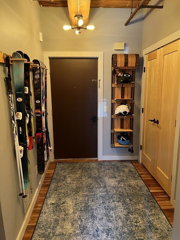 A hallway with a wooden door, skis and snowboards on the left, and a shelf with helmets and bags on the right.