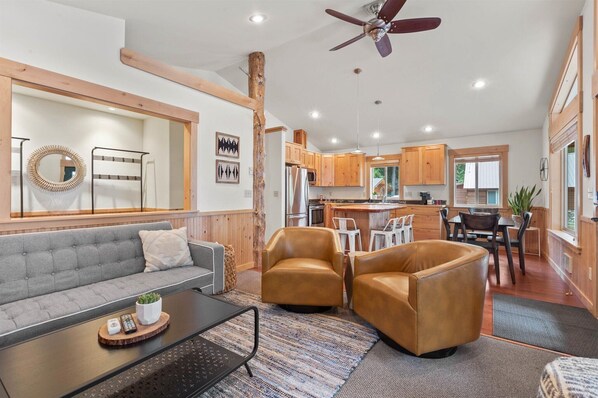 A modern living room with a grey sofa, two leather armchairs, a coffee table, and a dining area with a wooden table and chairs. - Priest Lake Family Cabin w/ Private Beach Access (Nordman)