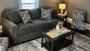 Una sala de estar con un sofá gris, cojines estampados, una mesa de centro con objetos decorativos, una mesita de noche con una lámpara y una silla con diseño.