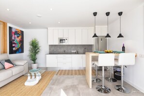 Una cocina moderna con armarios blancos, microondas integrado y un comedor con sillas blancas y una mesa de madera.