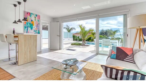 A modern living room with a glass coffee table, a sofa with patterned cushions, and a view of a pool and palm trees through large sliding glass doors. - TAJUYA SUITE (Los Llanos)