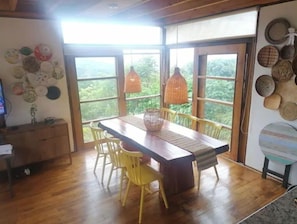 Un comedor con una mesa y sillas de madera, ventanas amplias con vistas a zonas verdes y adornos en la pared como cestas tejidas y platos.