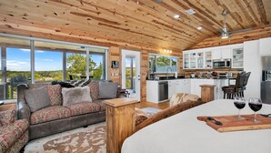 Un interior de cabaña acogedor con techo de madera, un sofá con cojines estampados, una zona de comedor con mesa de madera y una cocina con armarios blancos y electrodomésticos de acero inoxidable.