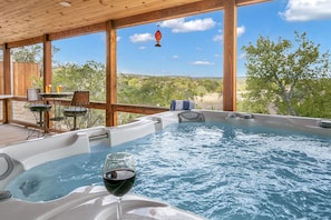 A hot tub with a glass of red wine, wooden deck, and outdoor seating area with a view of the landscape. - Spring Creek Casita at No Rancho Properties | Hot Tub | Hill Country Views & More (Fredericksburg)