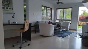 Ein modernes Wohnzimmer mit einer Couch, Sesseln, einem Couchtisch und einem Schreibtisch mit Stuhl. Große Fenster mit Jalousien, ein Deckenventilator und ein Poster an der Wand sind ebenfalls vorhanden.