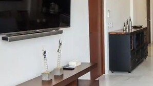 A modern living room with a large flat-screen TV mounted on the wall, a dark wooden entertainment unit below, and a kitchen area in the background. - CONDO LI BY TRVL2HM (Cabo San Lucas)