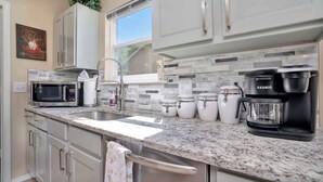 Cocina moderna con gabinetes blancos, encimera de granito, cafetera Keurig, microondas y ventana con vistas al exterior.