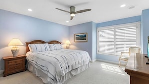 Un dormitorio con cama, mesitas de noche, escritorio, silla, ventilador de techo y ventana con persianas.