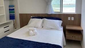 A neatly made bed with white and blue bedding, a wooden headboard, and a view of the beach through a large window. - Bela Hospedagem-Apt frente mar no Bessa (João Pessoa)