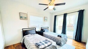 Quarto com duas camas, ventilador de teto e uma janela grande com cortinas.