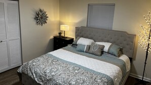 A bedroom with a large bed, a nightstand with a lamp, a decorative wall piece, and a window with blinds.