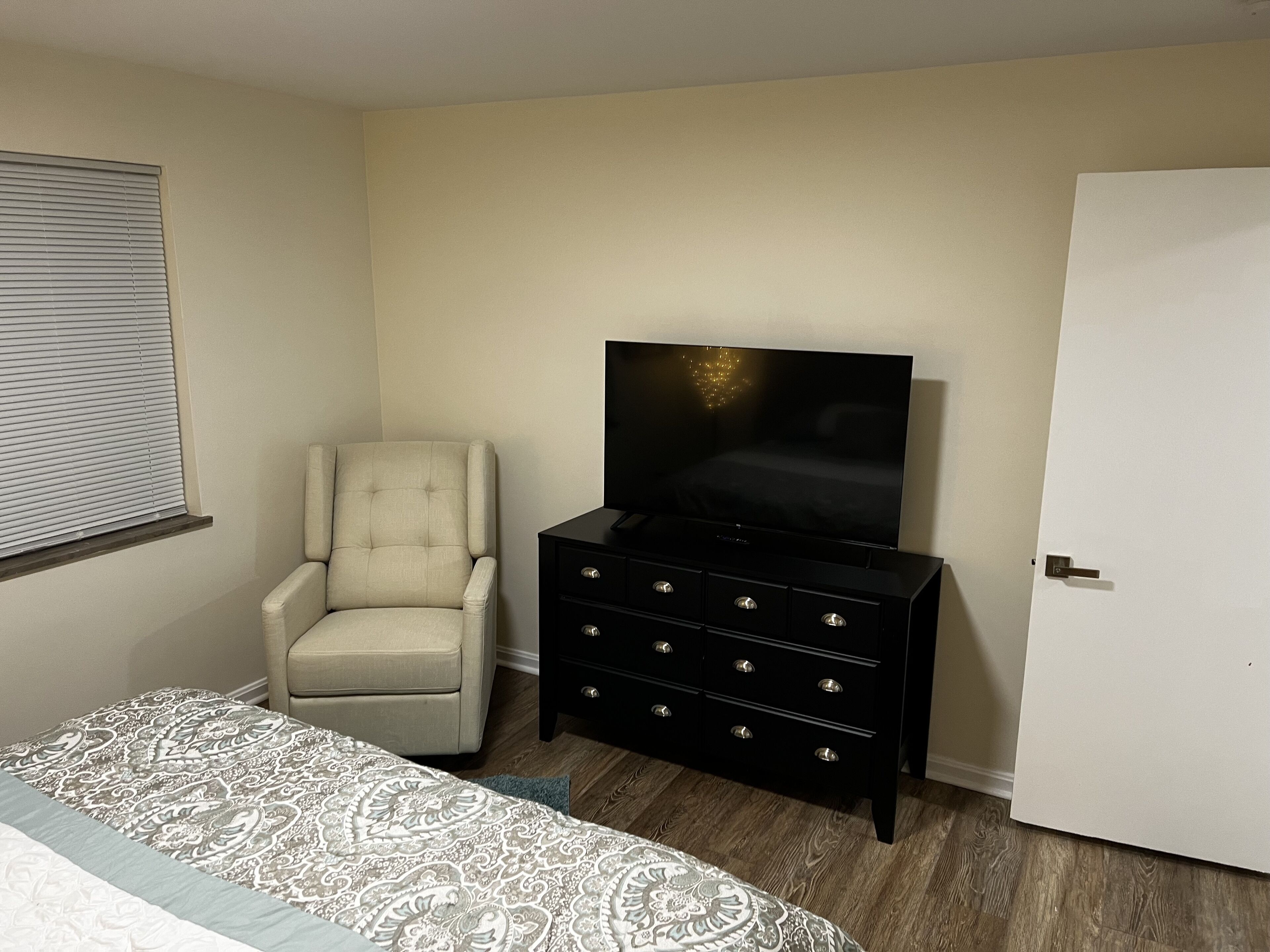 A bedroom with a bed, a chair, a TV on a dresser, and a door.