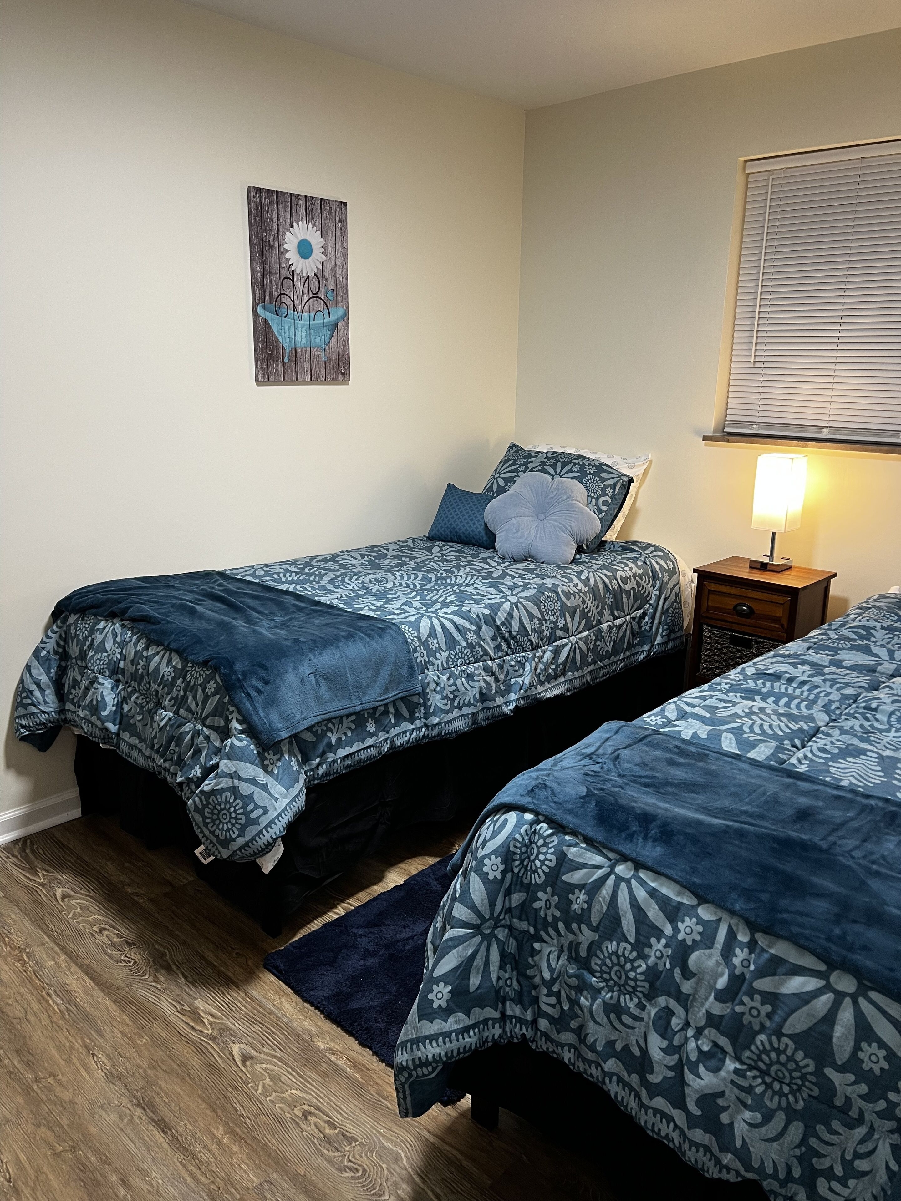 A bedroom with two beds, a wooden floor, a nightstand with a lamp, and a wall-mounted artwork.