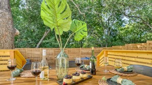 A wooden table set with wine glasses, bottles, and dessert plates in an outdoor setting with wooden benches and large green leaves in the background. - Bali Inspired | Ping-Pong | Yoga | Massage | Bocce (Austin)