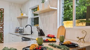 A modern kitchen with a large window, wooden shelves, and a variety of fruits and pastries on the counter. - Bali Inspired | Ping-Pong | Yoga | Massage | Bocce (Austin)